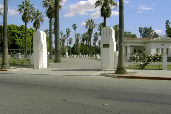 The entrance of Pomona Cemetery, showcasing its white Art Deco pillars, tall palm trees, and a welcoming pathway leading into the historic burial grounds.