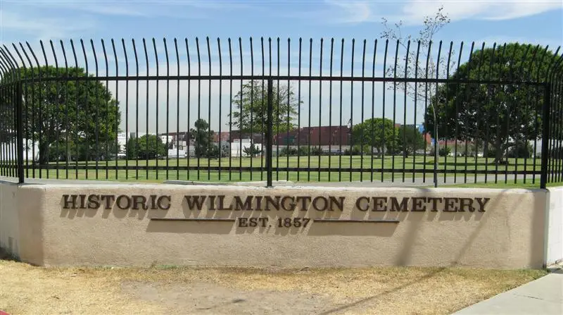 Historic entrance sign of Wilmington Cemetery in Los Angeles, CA, established in 1857, with a black iron fence and a well-maintained landscape.